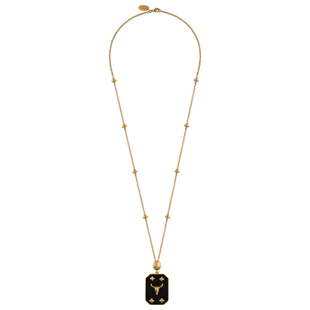 A gold necklace featuring a delicate chain with small star-like beads and a rectangular pendant with a bull symbol, displayed against a solid black background.